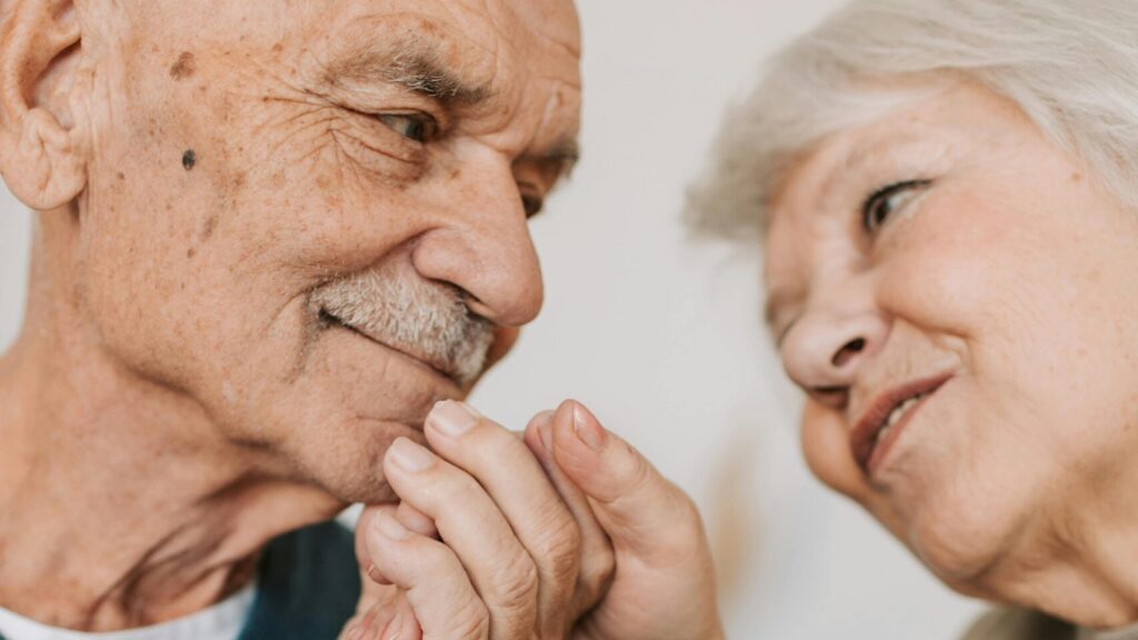 An elderly couple expressing love and affection, holding hands indoors.